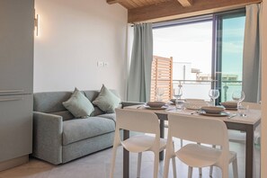 Ein modernes Wohnzimmer mit einem grauen Sofa, weißen Sesseln, einem Esstisch für zwei Personen und einem großen Fenster mit Vorhängen.