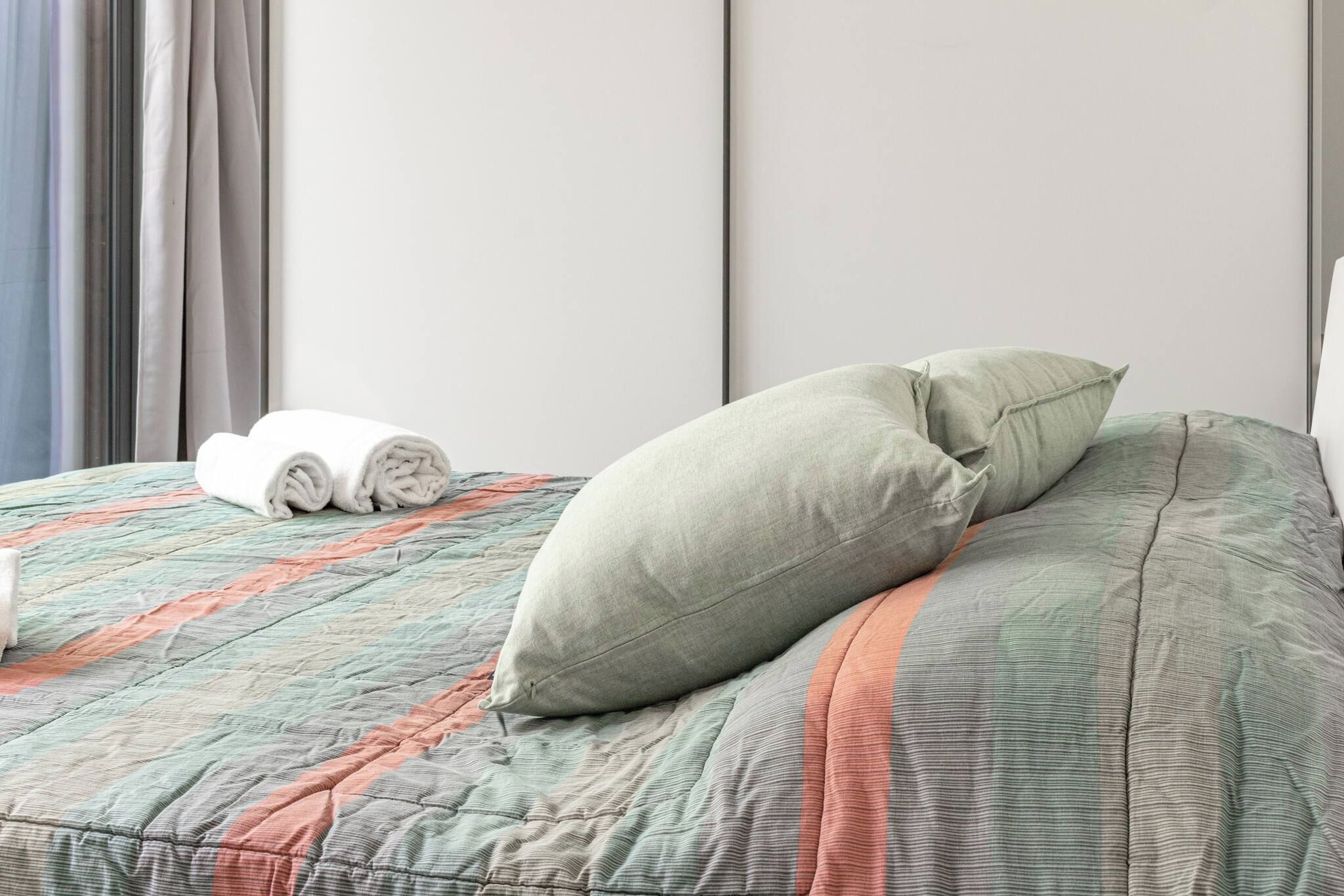 A neatly made bed with a quilted bedspread and two pillows, a rolled white towel, and a window with curtains.