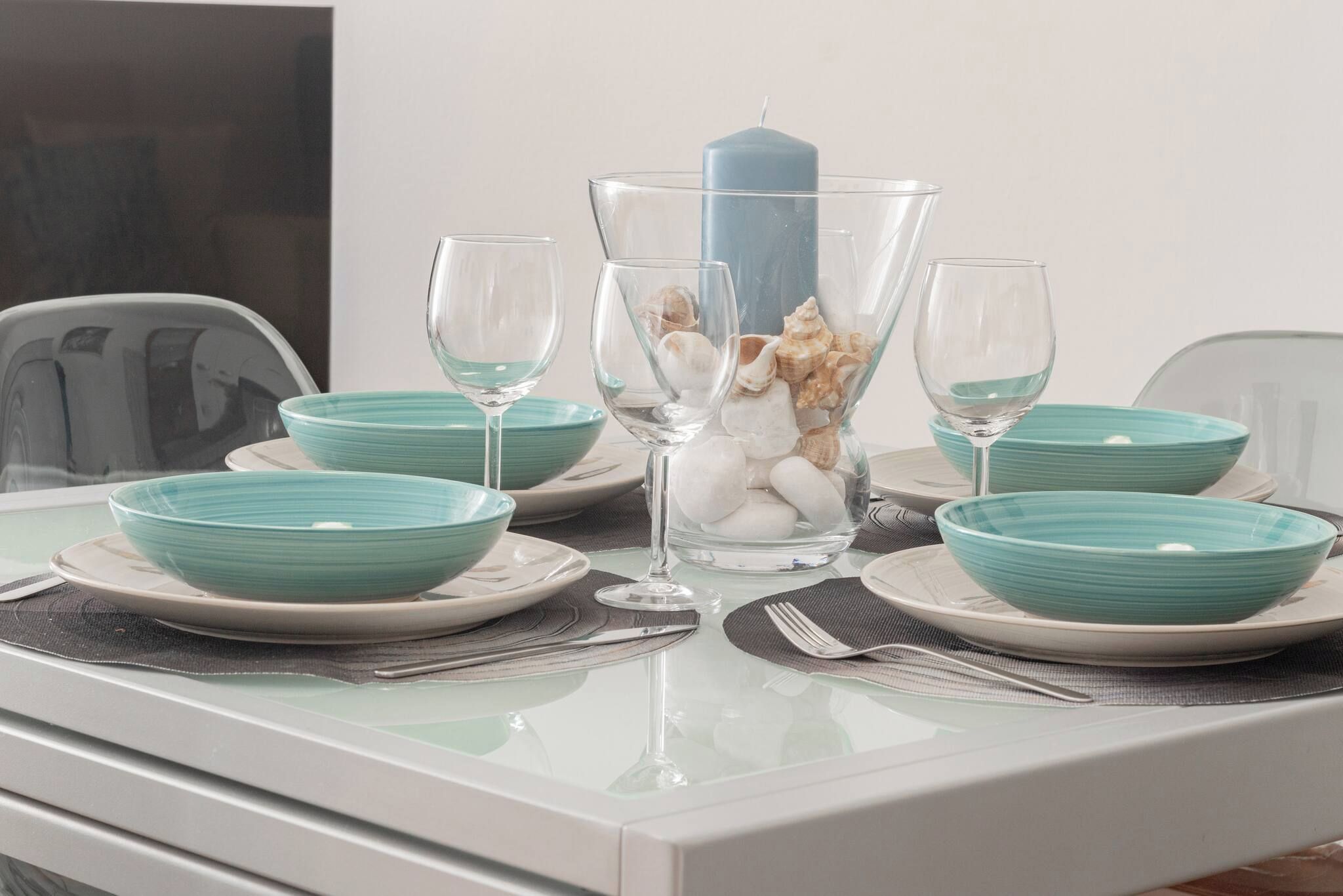 A dining table set with teal bowls, wine glasses, and a centerpiece with seashells and a candle.