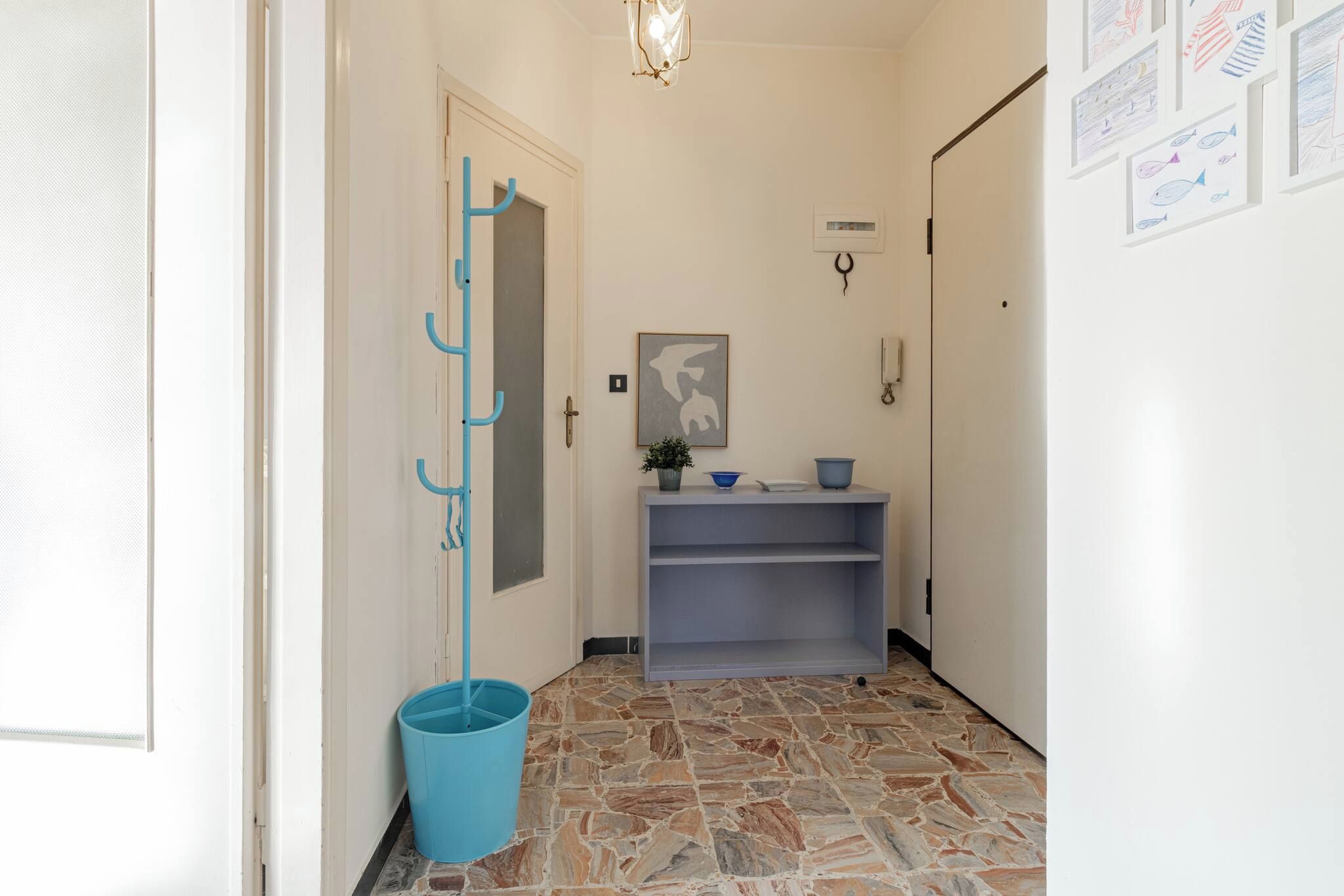 Un couloir avec un porte-manteau bleu, un seau bleu, une étagère grise et un sol imitant la pierre.