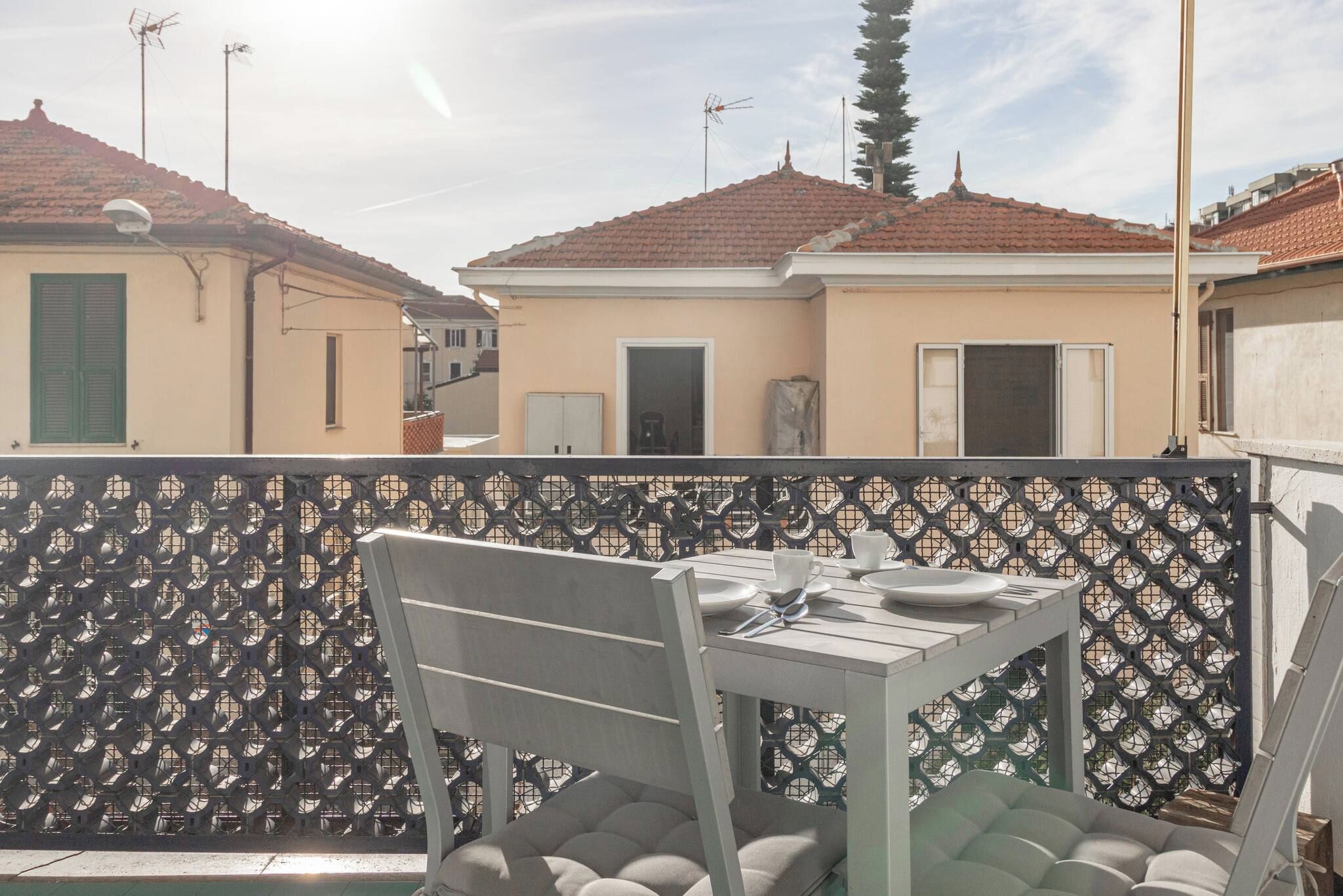 A balcony with a table and chairs, a patterned fence, and buildings with red-tiled roofs in the background.