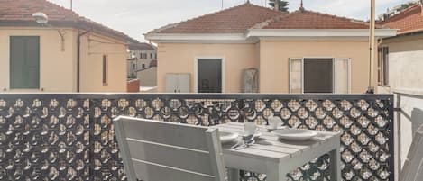 A balcony with a table and chairs, a patterned fence, and buildings with red-tiled roofs in the background.