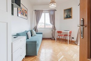 A compact living space with a sofa, a small table with chairs, a radiator, and a window with curtains. - APPARTAMENTO AZUL - Centro Sanremo (Sanremo)
