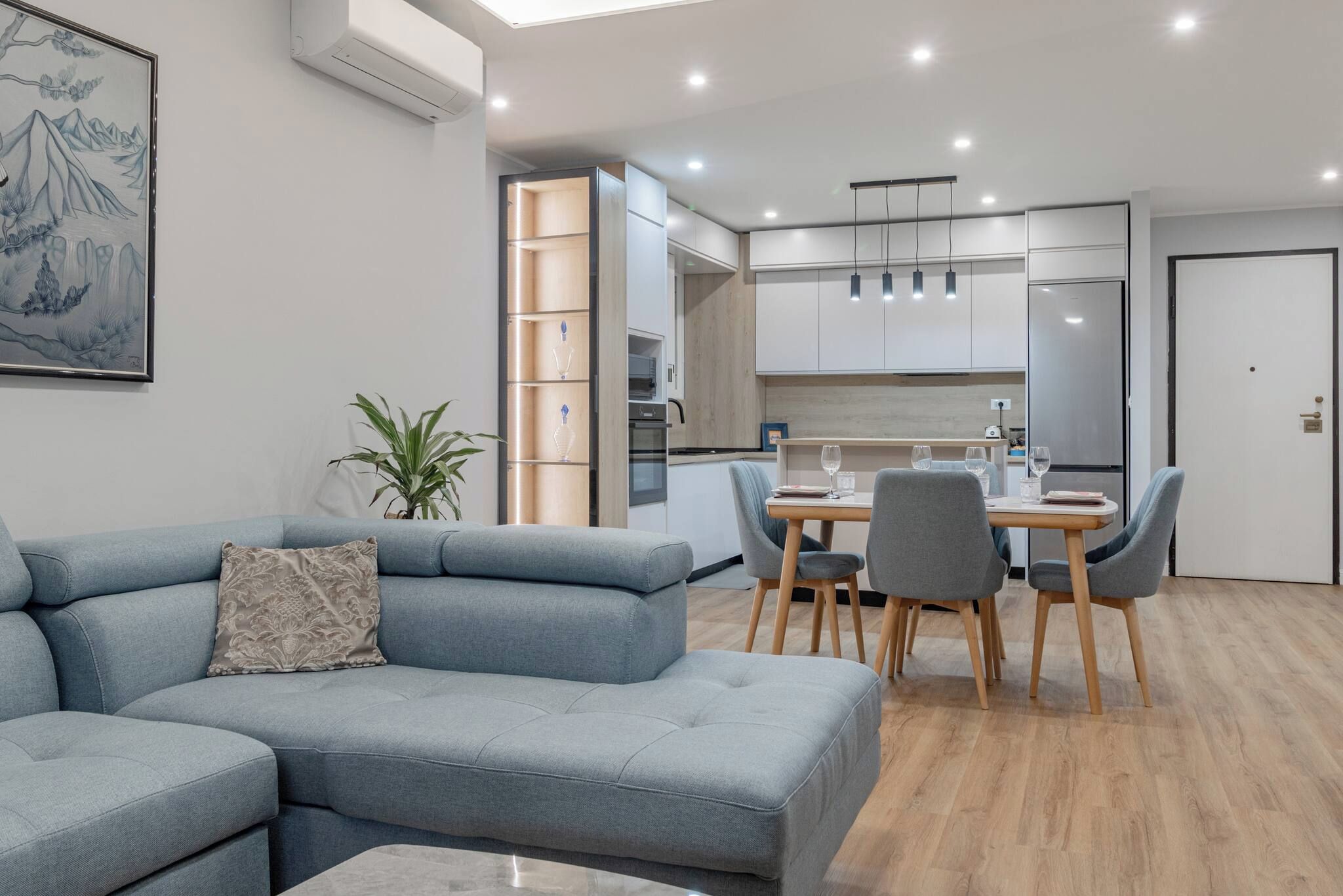A modern living room with a grey sectional sofa, a dining table with chairs, and a kitchen area with white cabinets and a refrigerator.