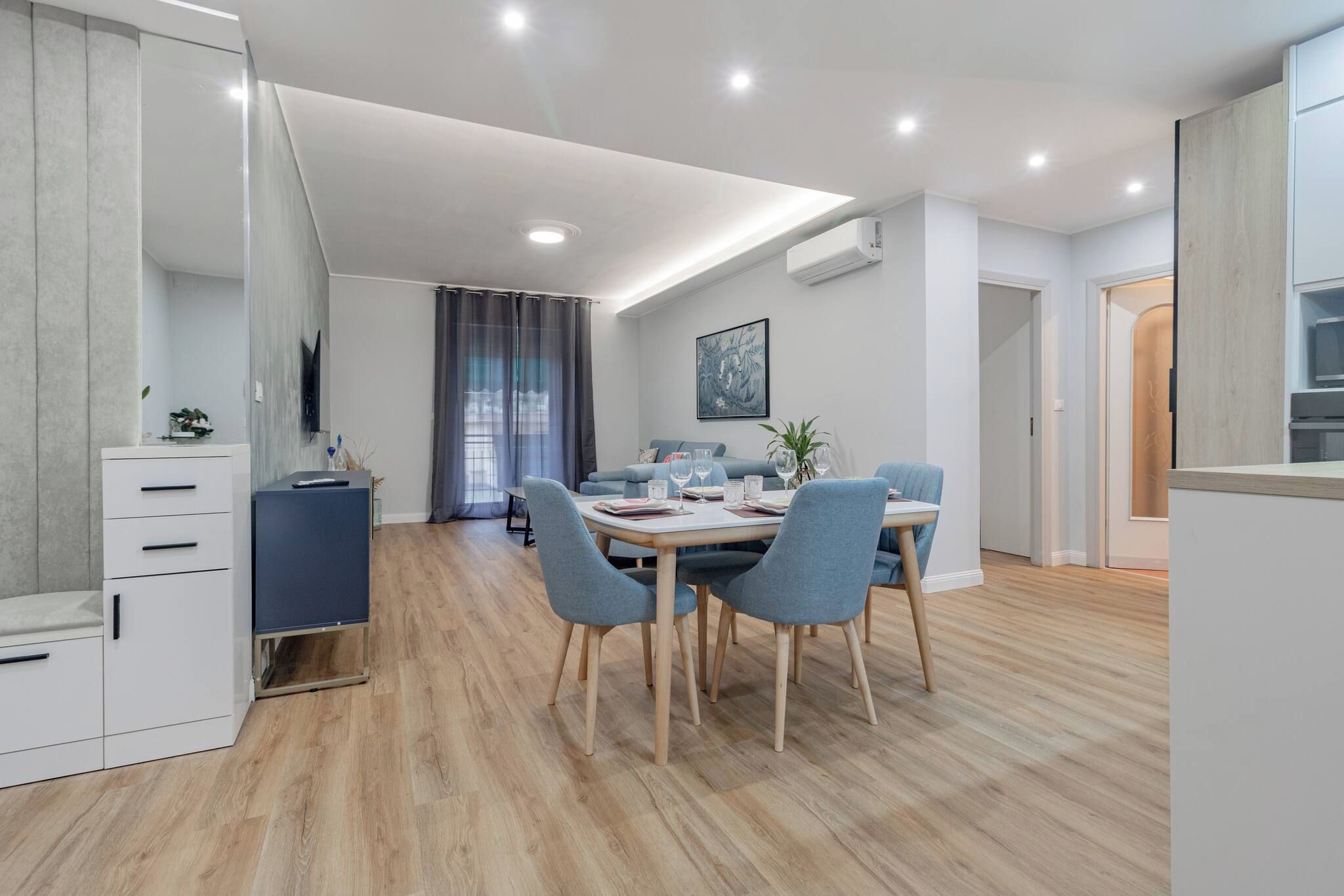 A modern kitchen with a dining area, wooden flooring, and a grey sofa.