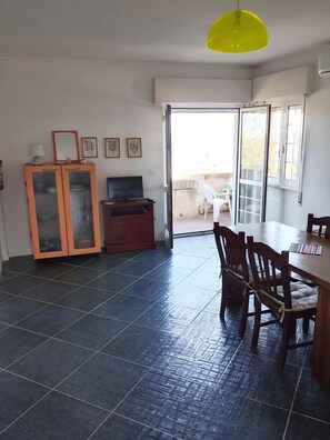 Ein Zimmer mit Esstisch, Stühlen, Fernseher, Schrank und einer Glastür zum Außenbereich.