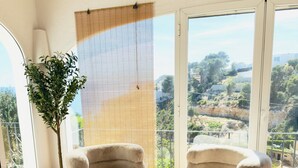 A room with two plush armchairs, a round wooden table, a large window with a view of a hilly landscape, and a hanging light fixture. - Villa Granadella with Sea Views in Jávea, Pool & BBQ for Weekly Retreats and Families (Costa Nova)