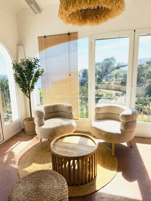 A room with two plush armchairs, a round wooden table, a large window with a view of a hilly landscape, and a hanging light fixture. - Villa Granadella with Sea Views in Jávea, Pool & BBQ for Weekly Retreats and Families (Costa Nova)