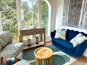 Ein Zimmer mit einem blauen Sofa, einem gemusterten Sessel, einem Holztischchen und einem großen Fenster mit Blick auf Bäume.