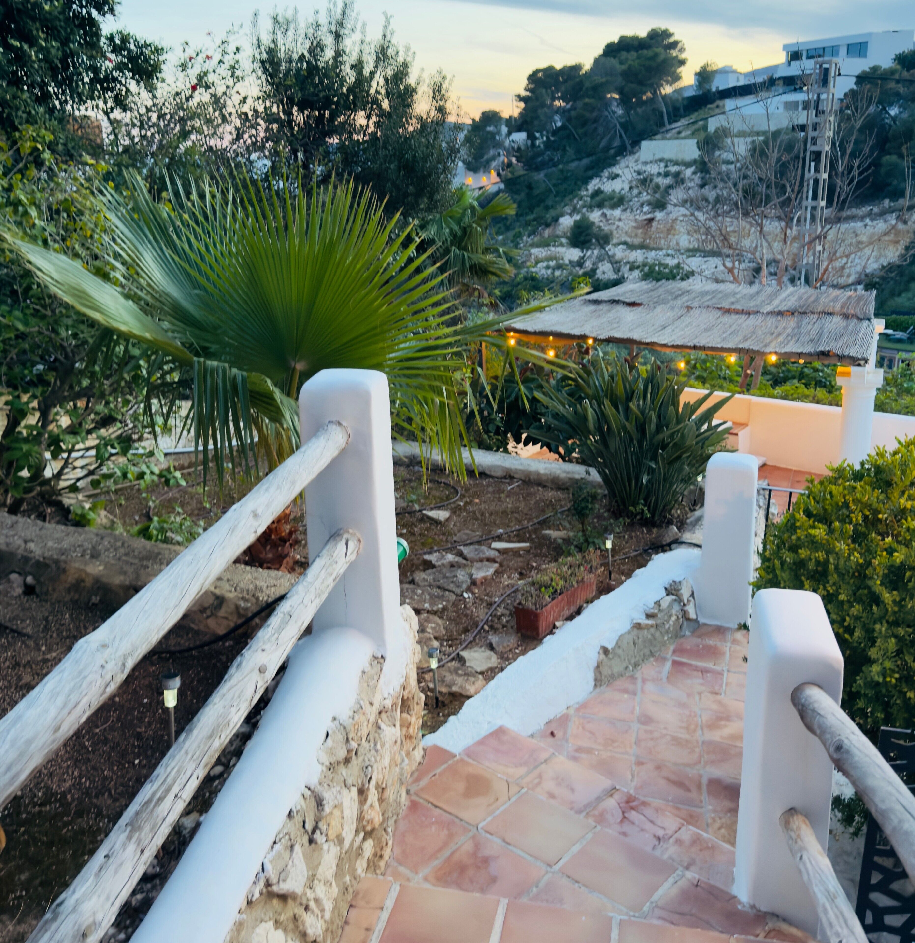 A staircase leading to a terrace with a thatched roof, surrounded by greenery and a view of a hillside.