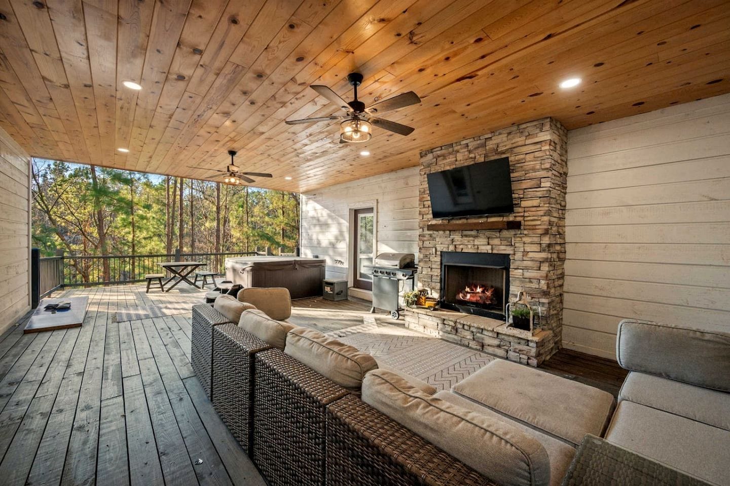 A covered deck with outdoor furniture, a stone fireplace, and a television.