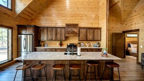 A spacious kitchen with wooden walls, a large island with bar stools, and a stainless steel refrigerator. - Cozy Cabin with Hot Tub & Fireplace in Broken Bow, Oklahoma (Broken Bow)