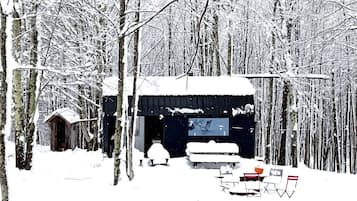Une cabane dans une forêt enneigée, avec un petit porche et une table entourée de chaises.