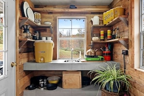 A compact kitchen with wooden walls, a window, and various kitchen appliances. - Off-Grid Tiny Cabin Retreat with Fire Pit & Forest Views in Bovina, New York (Bovina)
