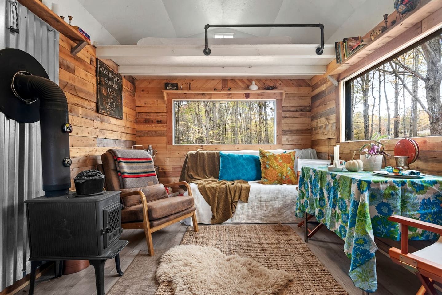 L’intérieur d’un chalet accueillant, avec un poêle à bois, un banc, une table recouverte d’une nappe à motifs floraux et une fenêtre donnant sur les arbres.