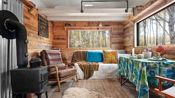 L’intérieur d’un chalet accueillant, avec un poêle à bois, un banc, une table recouverte d’une nappe à motifs floraux et une fenêtre donnant sur les arbres.