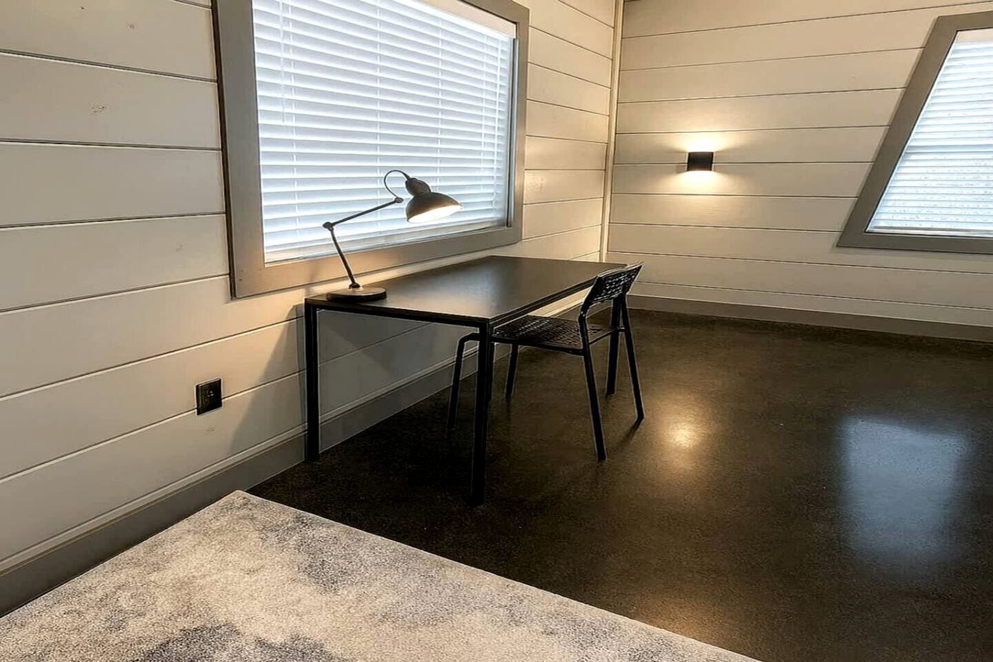 A room with a desk, chair, two windows with blinds, and a wall-mounted light fixture.