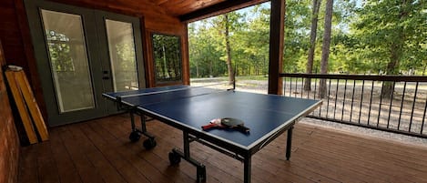 Mesa de ping pong sobre una terraza de madera con puertas de vidrio y vista a árboles.