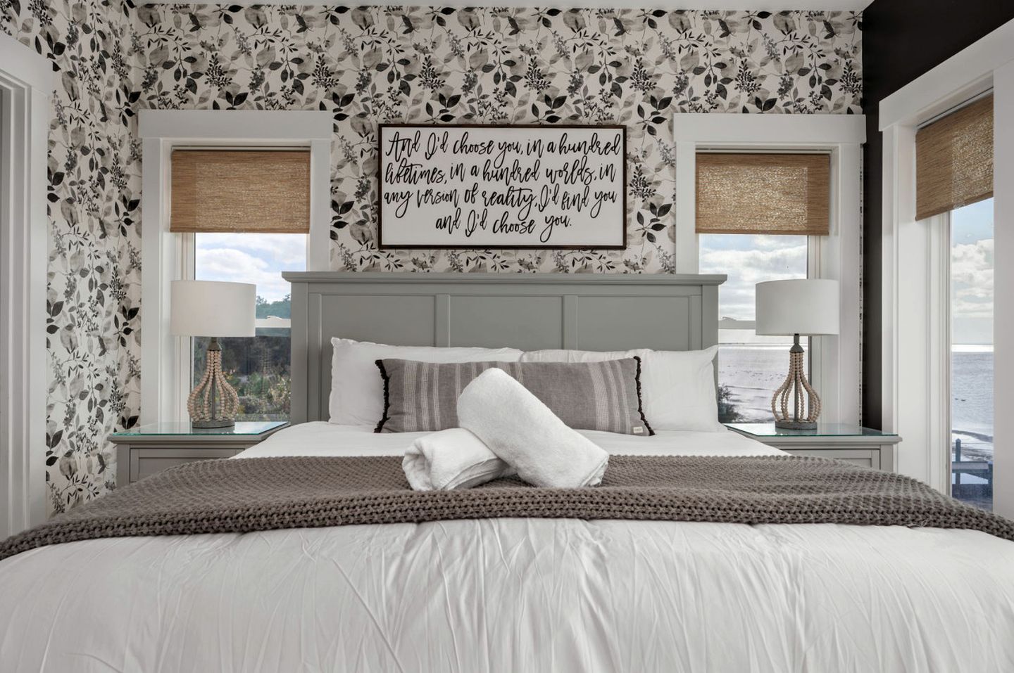A bedroom with a large bed, two bedside tables with lamps, a view of the sea, and a floral wallpaper.