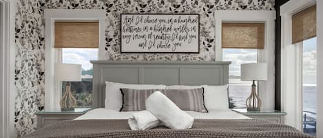 A bedroom with a large bed, two bedside tables with lamps, a view of the sea, and a floral wallpaper.
