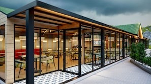 Una cafeteria moderna a l'aire lliure amb taules i cadires de fusta, parets de vidre i un paviment de rajoles.