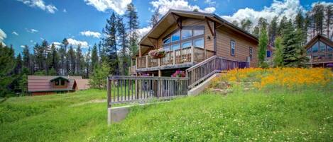 A two-story wooden cabin with a balcony, surrounded by a grassy field and a forest.