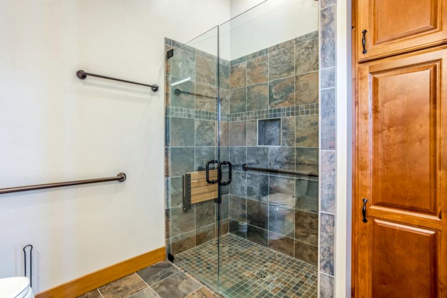 A glass-enclosed shower with a tiled floor, a wooden door, and a metal grab bar on the wall.