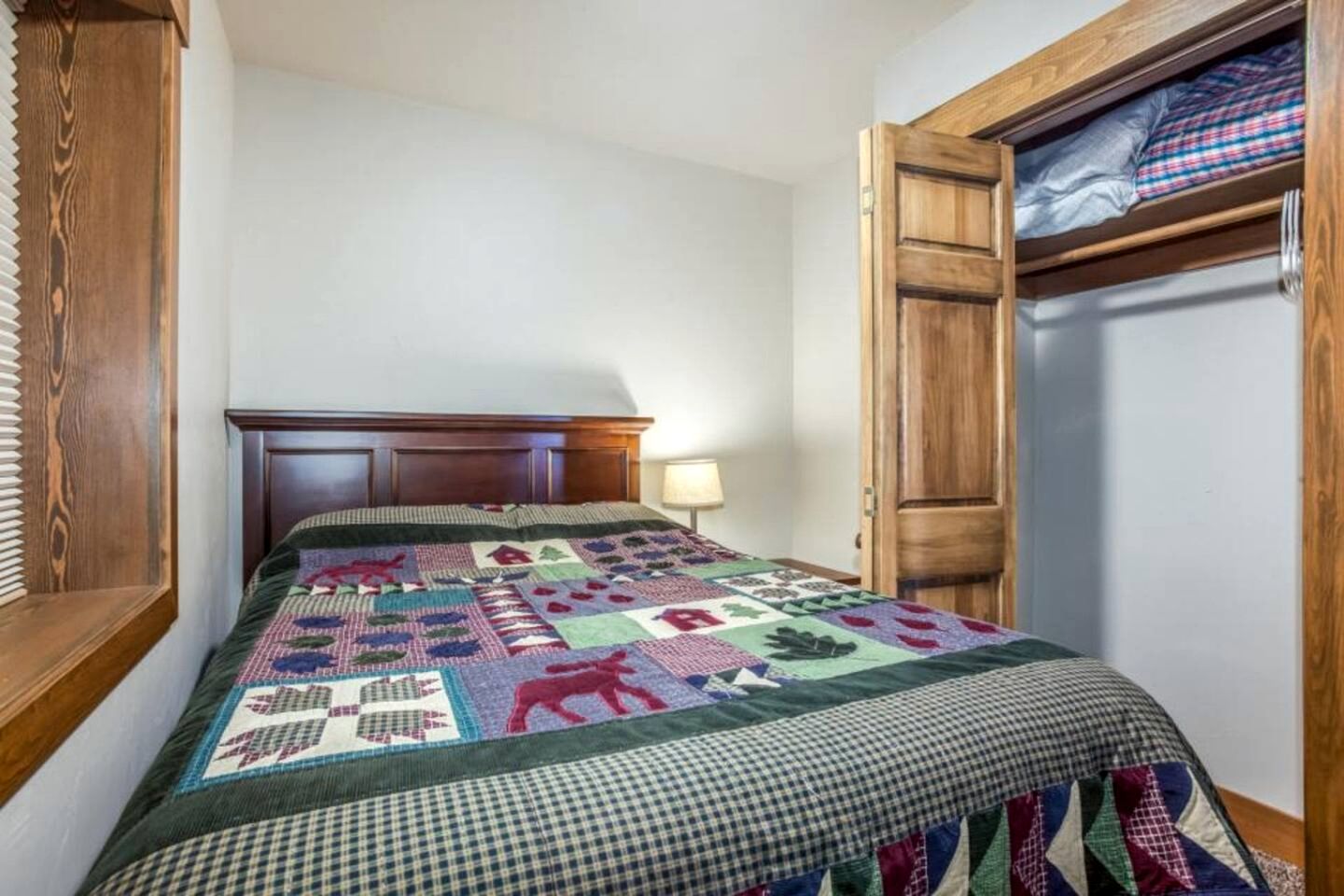 A bedroom with a wooden headboard, a quilt with various patterns, a bedside lamp, and a wooden wardrobe.