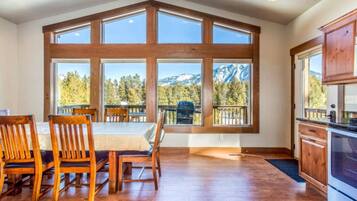 A dining area with a table and chairs, large windows with a mountain view, and a kitchen with wooden cabinets.