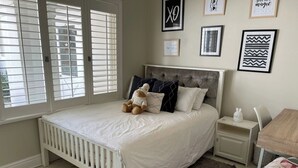 Um quarto com cama branca, um urso de pelúcia, uma mesa de cabeceira, uma cadeira e uma escrivaninha com uma luminária.