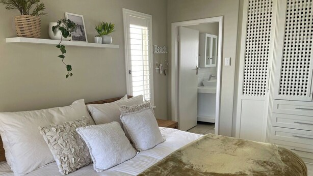 Quarto com cama, almofadas, mesa de cabeceira, planta pendente, banheiro visível por uma porta aberta e guarda-roupa branco.