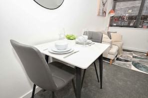 A dining area with a white table set for two, a grey chair, and a sofa in the background.