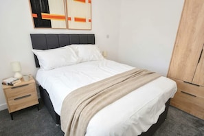 Ein ordentlich bezogenes Bett mit Kopfteil, ein Nachttisch mit Lampe und ein Kleiderschrank.