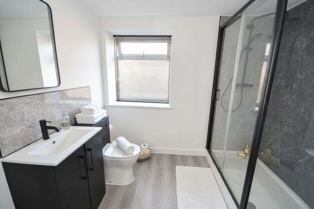 Ein modernes Badezimmer mit einem weißen Waschbecken, dunklen Schränken, einer Toilette, einer Duschkabine mit Glastür und einem Fenster mit Jalousien.