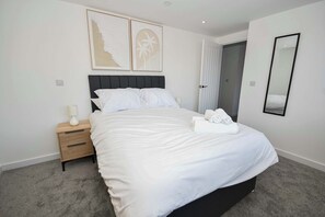 Een net opgemaakt bed met witte lakens, een nachtkastje met een lamp en een spiegel aan de muur.