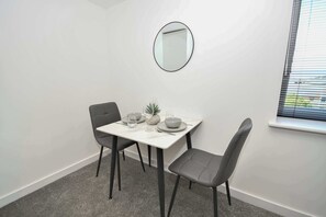 Een gezellig eetgedeelte met een witte tafel, twee grijze stoelen, een ronde spiegel aan de muur en een raam met zonwering.