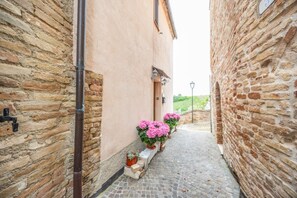 Ein schmaler Gang mit Steinmauern, einem gepflasterten Weg und Kübeln mit rosa Blüten.