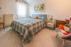 Ein Schlafzimmer mit Bett, Nachttisch, Sessel, Schreibtisch, Heizkörper und einem Fenster mit Vorhängen.