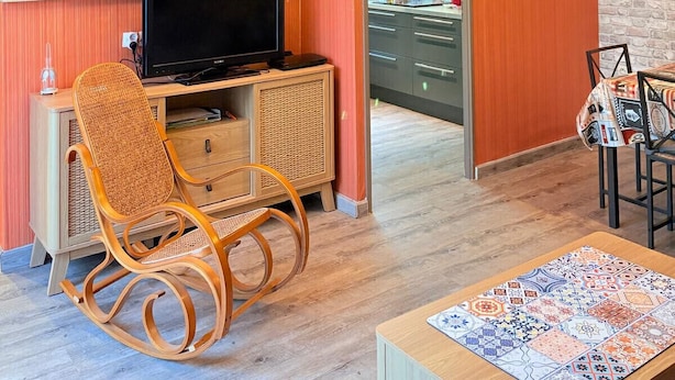 Una sala de estar con televisor, una mecedora, un comedor con mesa, y una cocina visible a través de una puerta abierta.