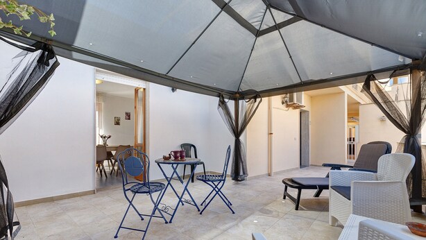 Un espace extérieur abrité, aménagé avec des chaises et une table, doté d’un sol carrelé et d’une porte-fenêtre donnant sur un espace intérieur.