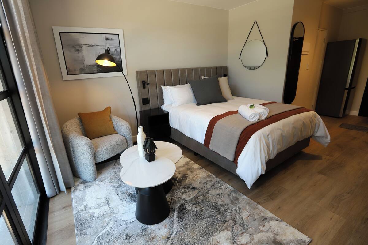 A hotel room with a bed, bedside table, lamp, chair, and a painting on the wall.