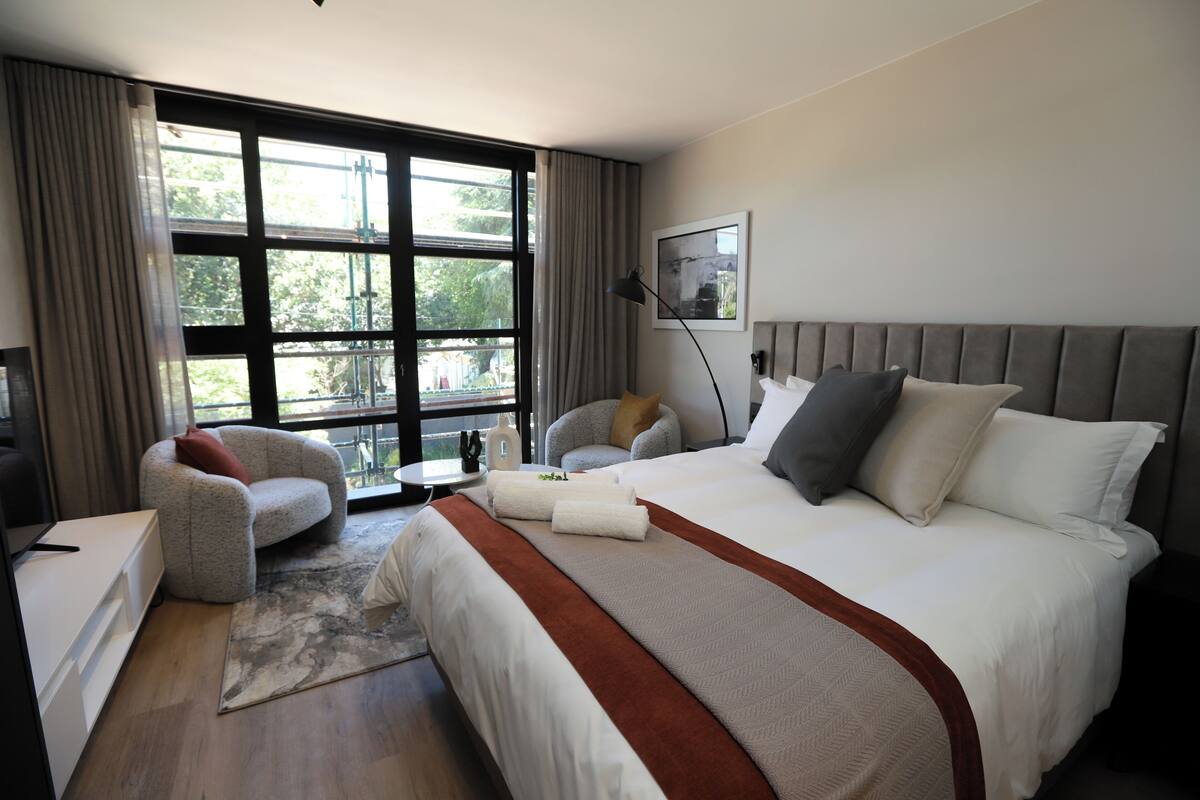 A hotel room with a large bed, a sofa, a desk, and a view of the outdoors through a large window.