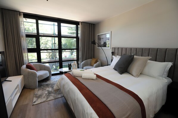 A hotel room with a large bed, a sofa, a desk, and a view of the outdoors through a large window.