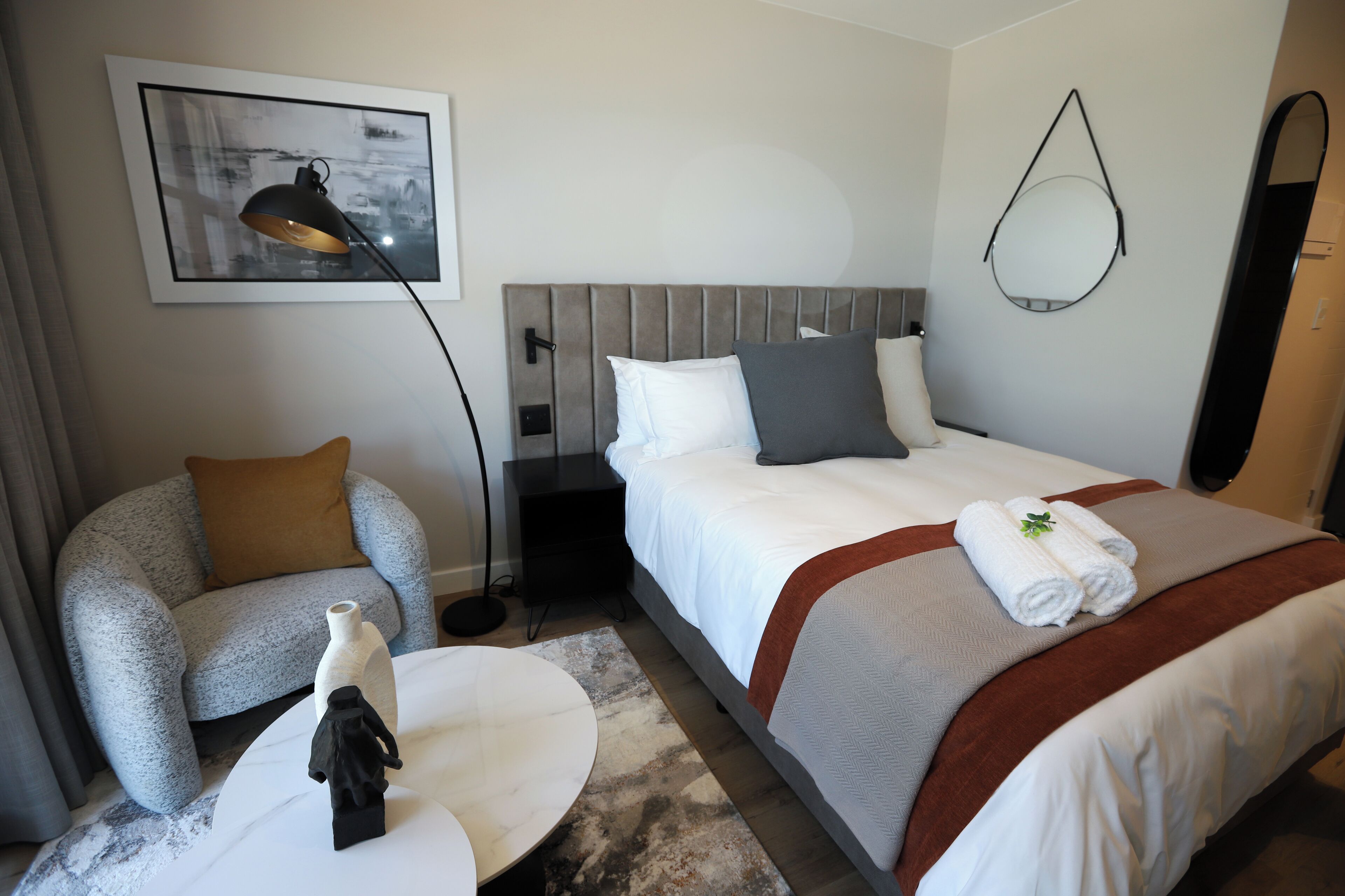 A hotel room with a bed, a chair, a small table, a lamp, and a wall-mounted mirror.