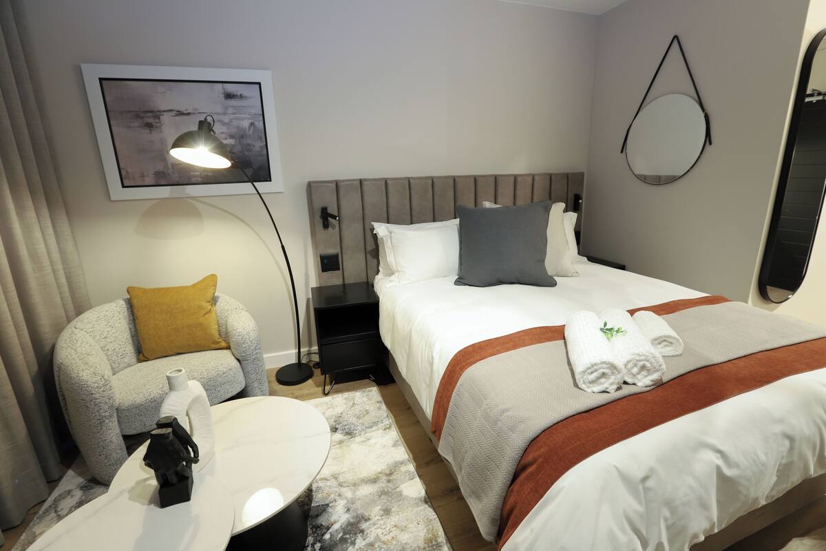 A hotel room with a bed, a chair, a table, a lamp, a painting, and a mirror.
