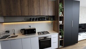 Una cocina moderna con armarios blancos, horno empotrado, cocina a gas y microondas. Hay una repisa de madera con objetos decorativos y una planta en maceta.