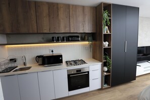 A modern kitchen with white cabinets, a built-in oven, a gas stove, and a microwave. There is a wooden shelf with decorative items and a potted plant. - WINK Aparthotel Saxon Square (Johannesburg)