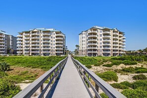 Ein hölzerner Steg führt zu einem Strandhotel mit mehreren Balkonen und blauem Himmel.