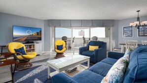 Una sala de estar moderna con un sofá azul, sillones amarillos, un televisor de pantalla plana y un ventanal con vistas a la ciudad.