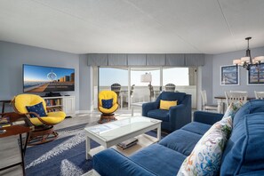Un salon moderne avec un canapé bleu, des fauteuils jaunes, un téléviseur à écran plat et une grande fenêtre donnant sur la ville.